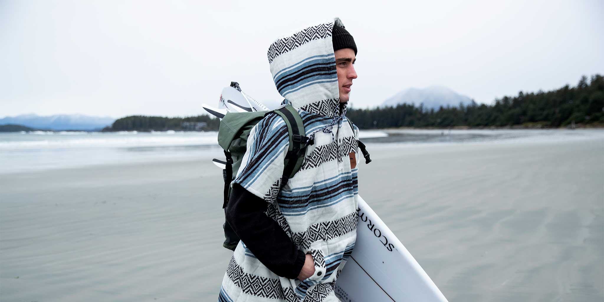 A person in a hooded poncho and beanie stands on a sandy beach, holding a surfboard and wearing a backpack, with trees and mountains in the background.