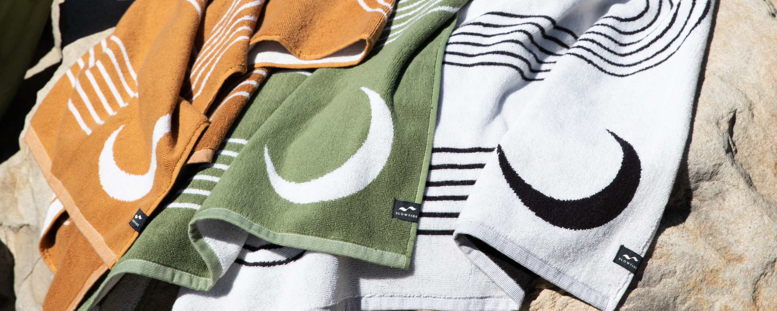 Three towels in burnt orange, olive green, and white with crescent moon and stripe designs are draped over a large rock in sunlight.