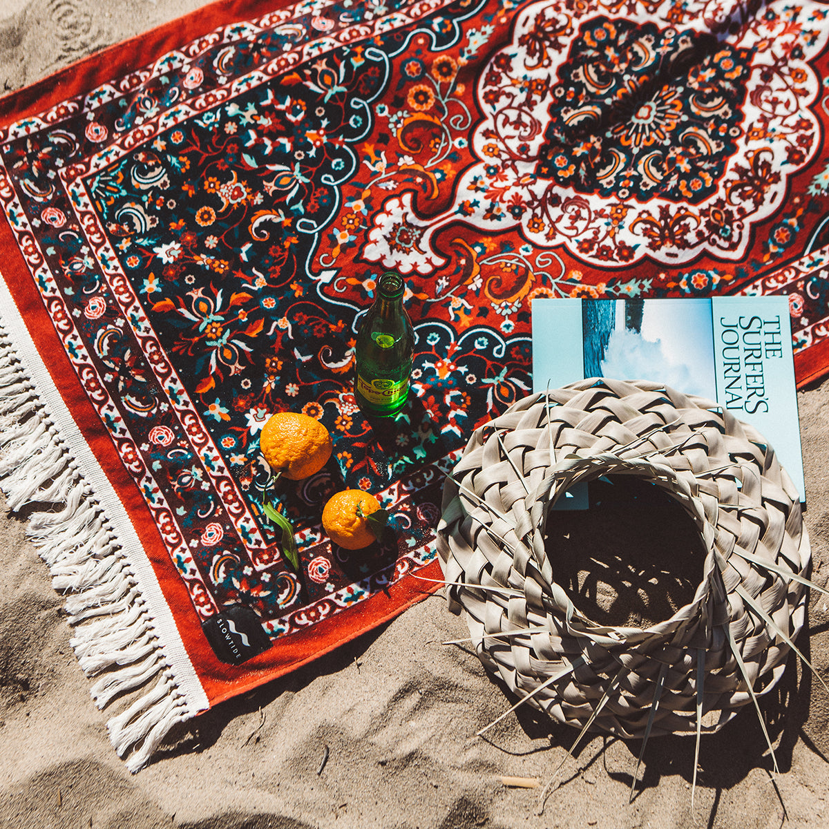 A patterned beach towel on sand with two oranges, a green glass bottle, a magazine, and a woven sun hat. Sunlight casts shadows on the colorful towel.