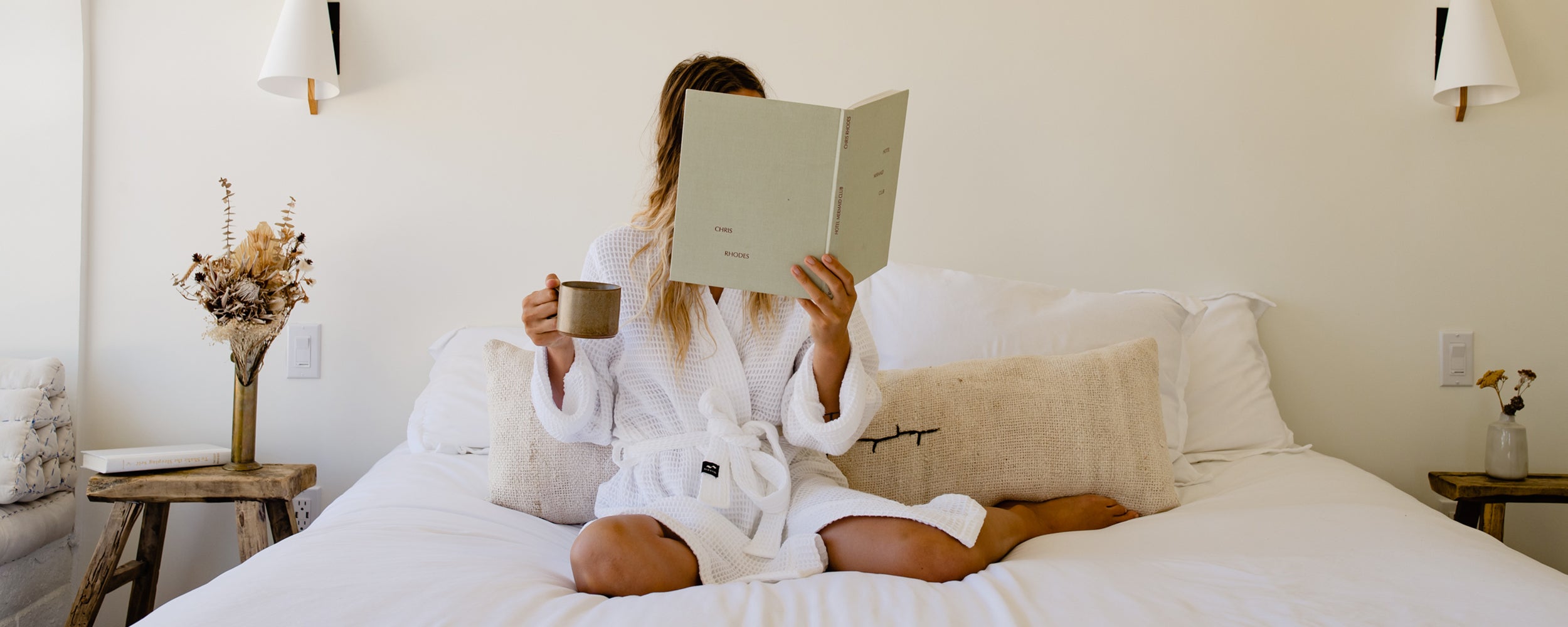 A person in a white bathrobe sits cross-legged on a bed, holding a mug in one hand and reading a book or menu. The room is decorated in neutral tones with dried flowers on a wooden nightstand.