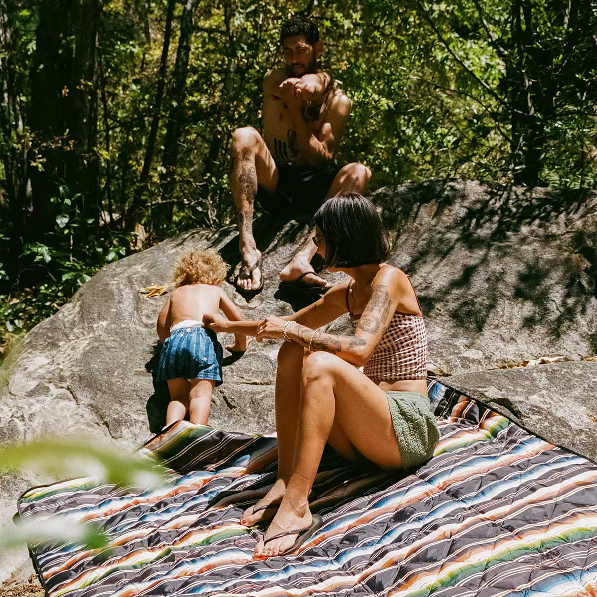 A woman sits on a striped blanket outdoors, a toddler in blue shorts climbs a large rock, and a man sits shirtless on the rock surrounded by trees in a sunny, wooded area.