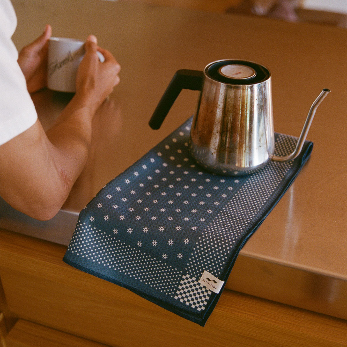 A person holds a mug at a counter, next to a metal gooseneck kettle resting on a patterned blue towel.