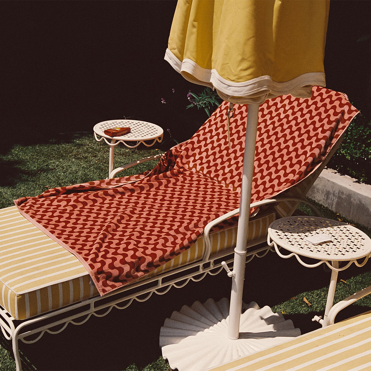 A lounge chair with a red wavy-patterned towel and yellow-striped cushion sits under a yellow umbrella, next to two small white tables in a sunny, grassy outdoor area.