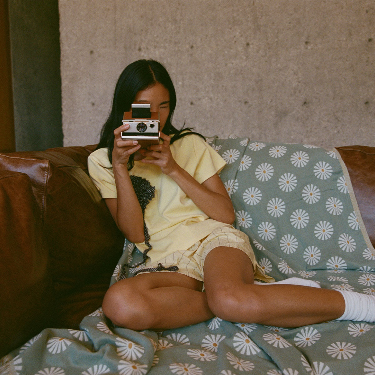 A woman sits on a brown sofa covered with a green daisy-patterned blanket, holding a vintage instant camera up to her face and wearing a yellow outfit with white socks.