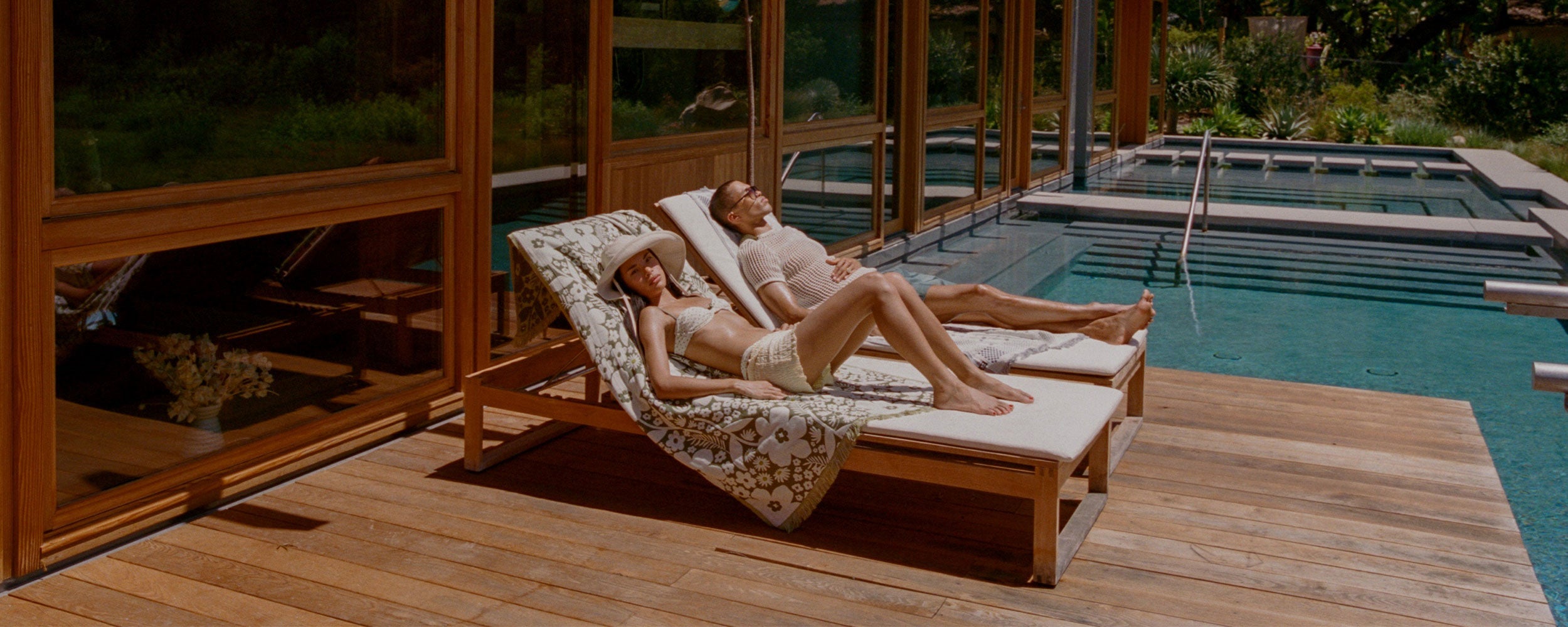 Two people relax on lounge chairs by a modern pool, sunbathing on a wooden deck beside a glass-walled building, with trees and greenery reflected in the windows.