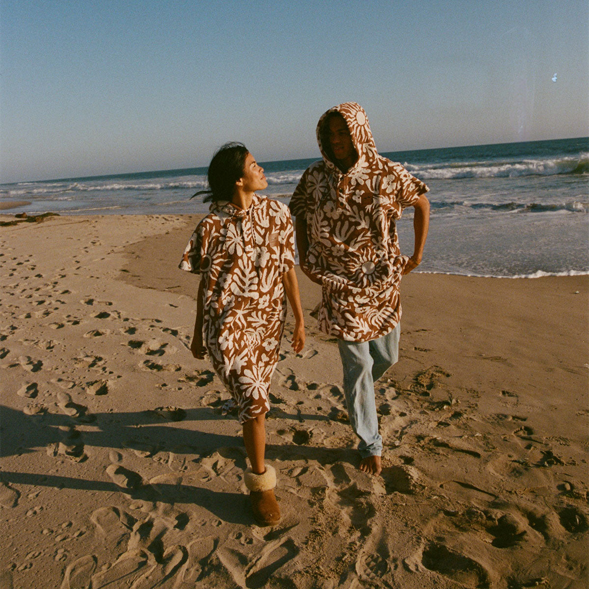 Two people walk along a sandy beach at sunset, both wearing matching brown and white floral hooded outfits. One person looks at the other as ocean waves roll in behind them.