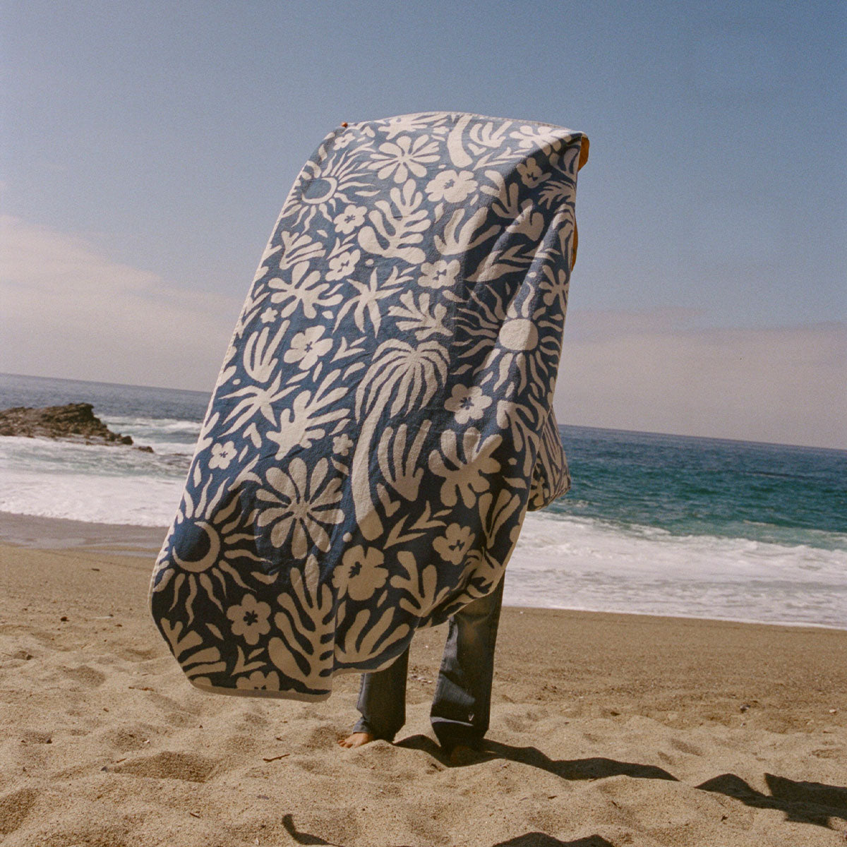 A person stands on a sandy beach, wrapped in a large blue and white floral-patterned blanket, facing the ocean with waves in the background under a clear sky.