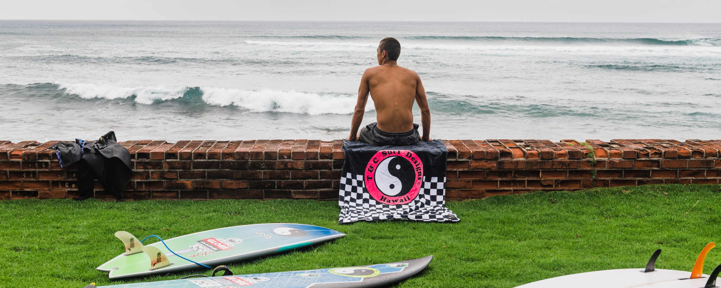 A shirtless man sits on a brick wall overlooking the ocean, with a Hawaiian Island Creations towel beneath him and several surfboards lying on the grass nearby. Waves roll in under a cloudy sky.