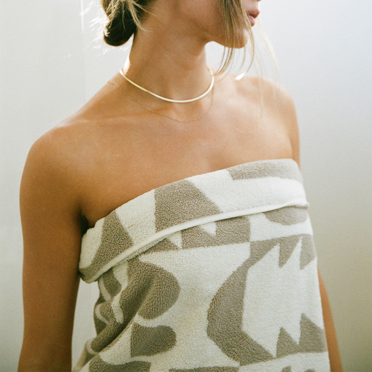 A woman with light hair wears a gold necklace and a beige and white patterned towel wrapped around her chest, standing in soft lighting. Her face is partially out of frame.