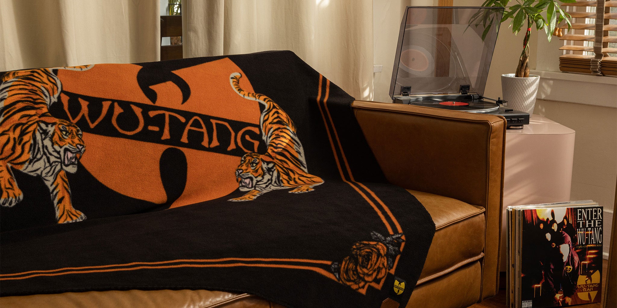 A brown couch with a Wu-Tang Clan blanket featuring tigers draped over it, next to a turntable and a vinyl record of Enter the Wu-Tang (36 Chambers) in a cozy living room.