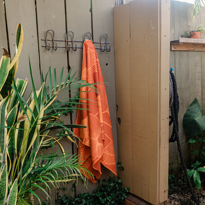 Towel with Calathea leaf pattern hanging on wooden fence outdoors near plants and shower.