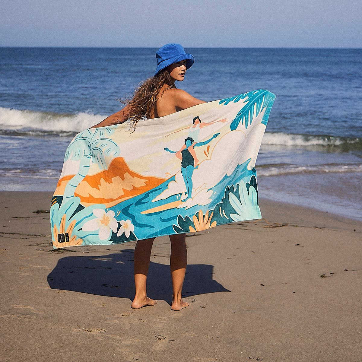 Cotton beach towel, absorbent, displayed with blue hat on sandy shore.