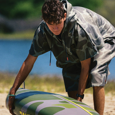Camo surf changing poncho with hood, on sandy beach near water.