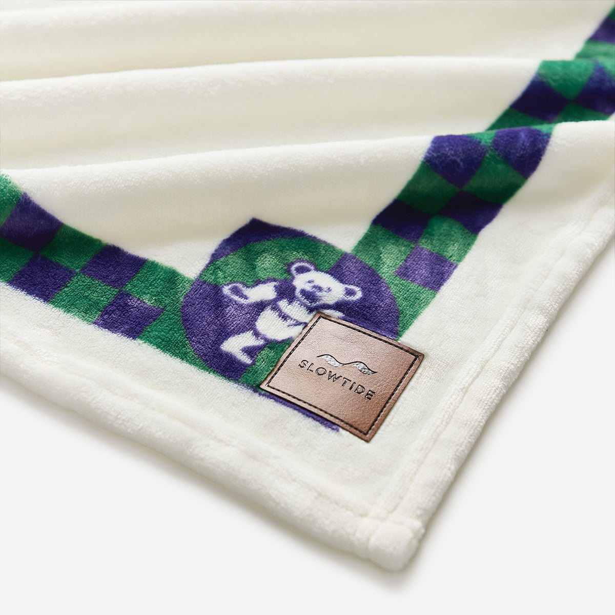 Oversized plush throw blanket with green and purple bear pattern and small brown logo patch.