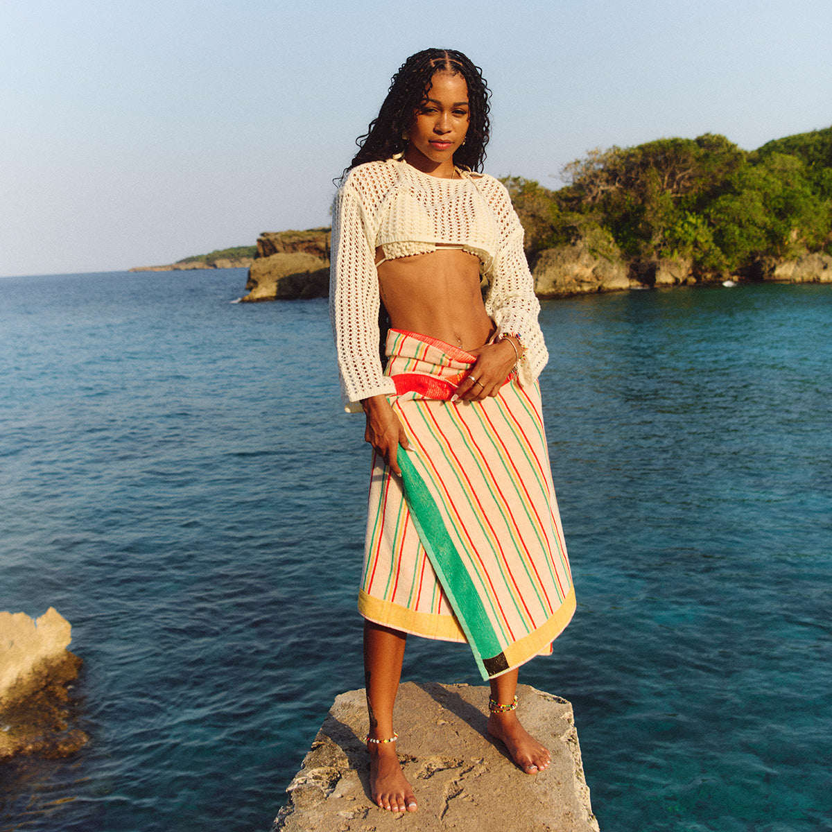 Crochet top and striped skirt with sustainable cotton beach towel, coastal setting.