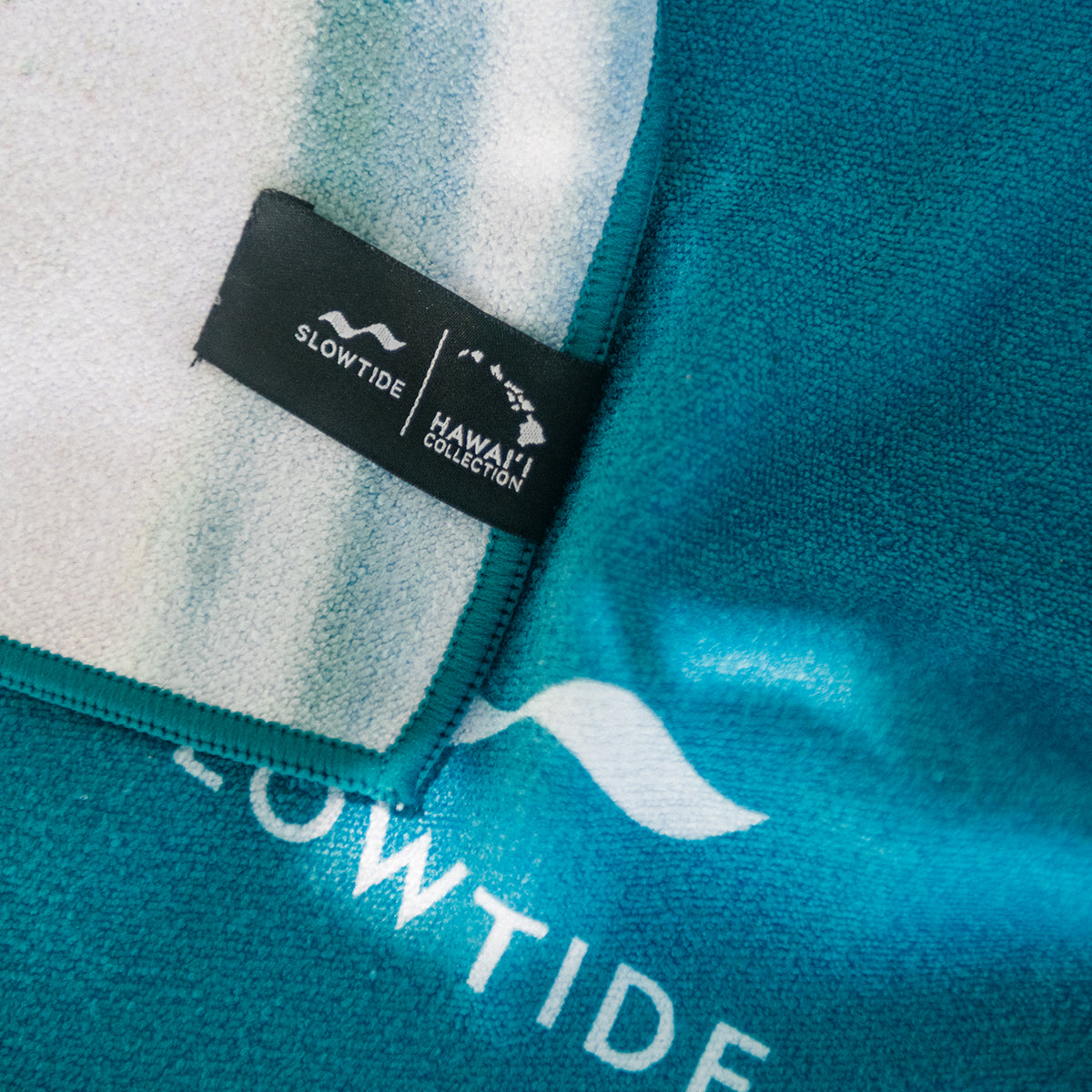 Teal quick-dry towel with white wave logo and small collection tag.