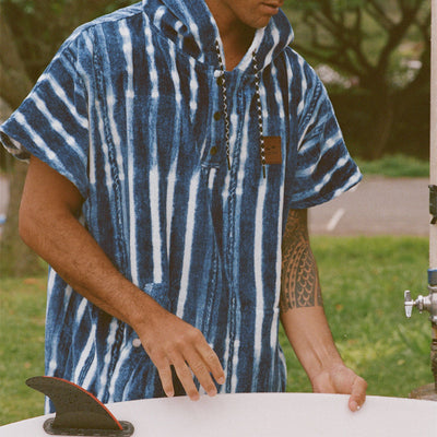 Cotton poncho with blue stripes, absorbent design, shown near a surfboard outdoors.