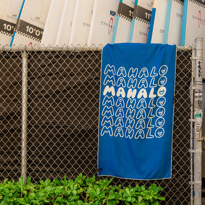 Blue quick-drying towel with text print hanging by surfboards and plants.