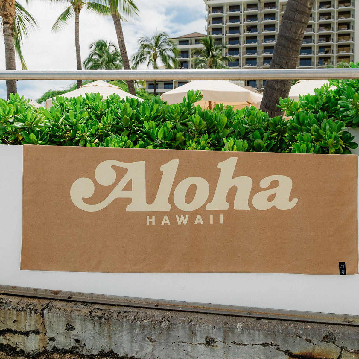 Beige quick-drying towel with Aloha Hawaii text, displayed hanging against a simple backdrop.