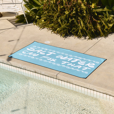 Blue quick-drying towel with Salt Water Can Fix That text, poolside.