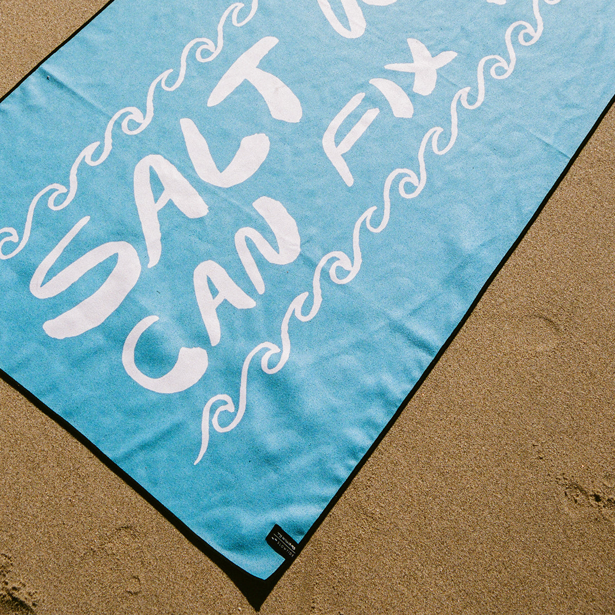 Blue sustainable travel towel with white wavy text on sand.