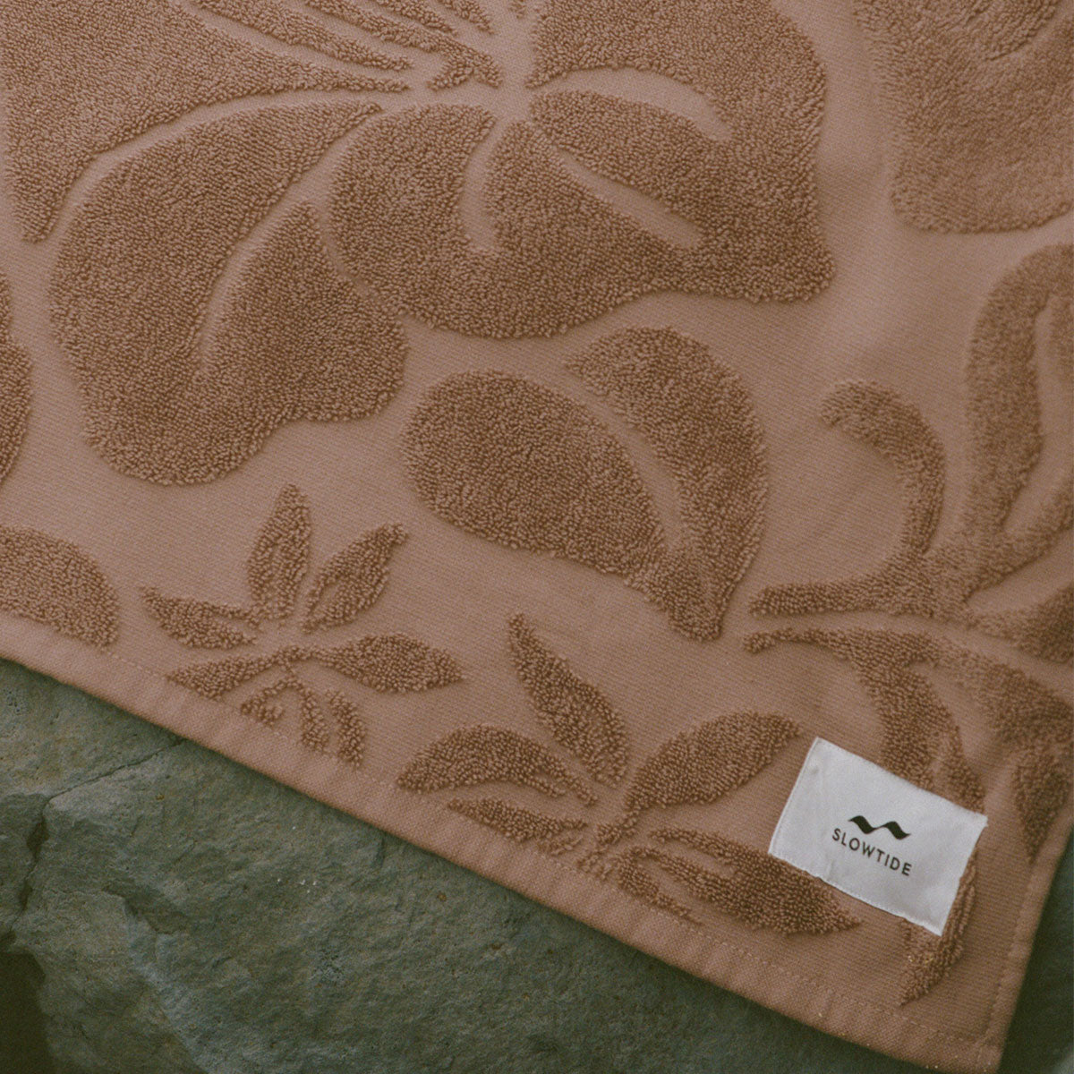 Brown cotton beach towel with floral pattern, absorbent, displayed on green stone.