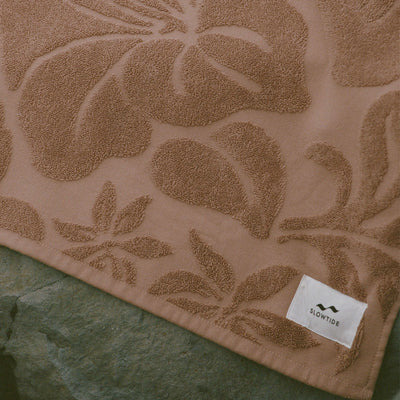 Brown cotton beach towel with floral pattern, absorbent, displayed on green stone.