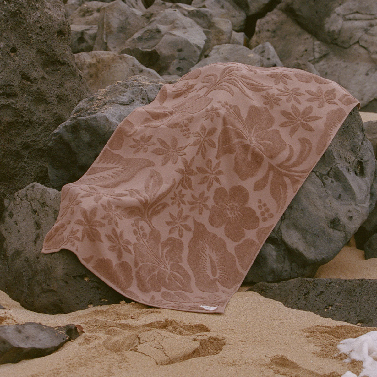 Beach towel, absorbent material, brown floral print, displayed on rocks on sand.
