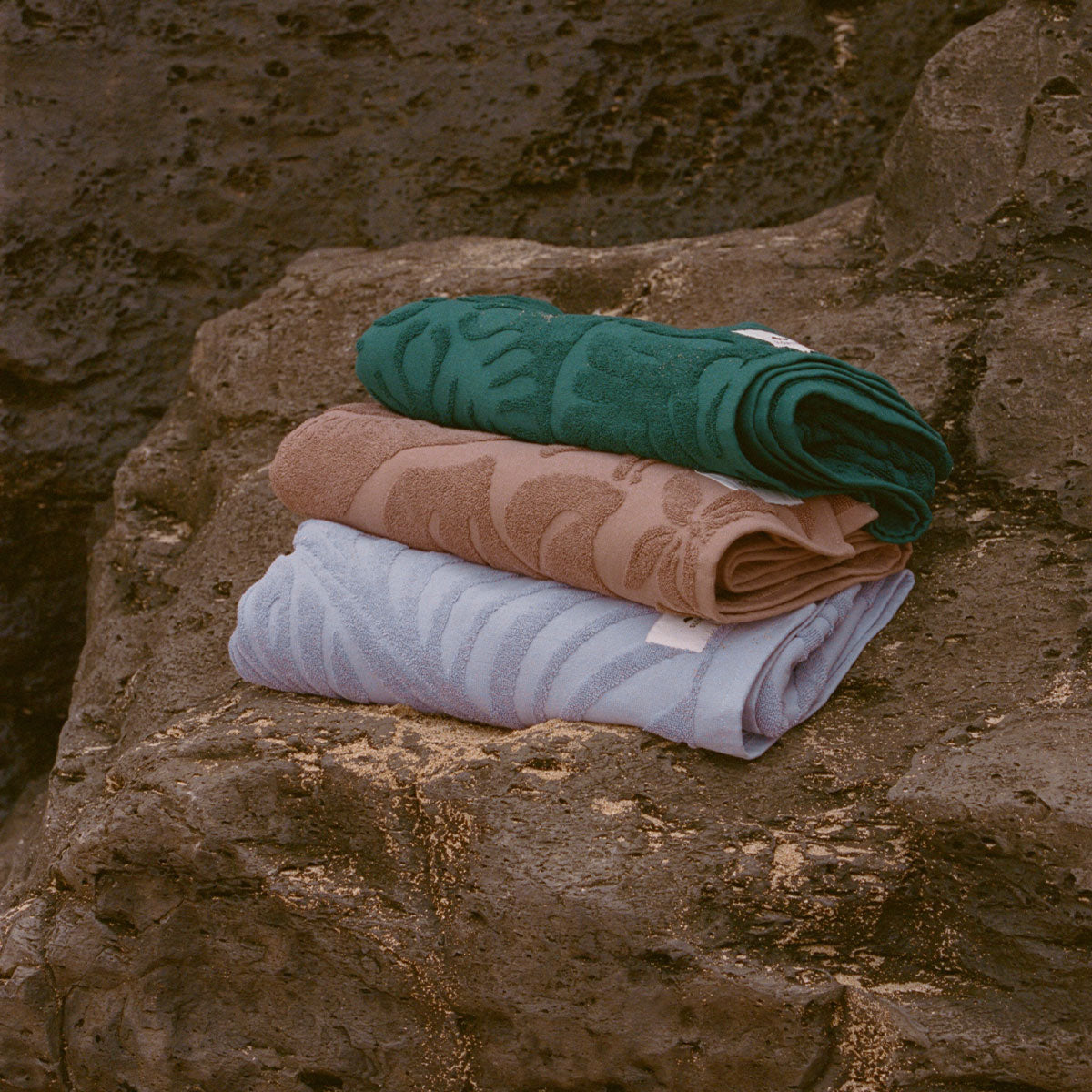 Velour towels set in green, brown, and blue stacked on a textured rock surface.