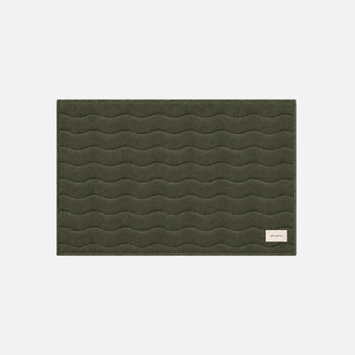Cotton bath mat in dark green with wavy stitching and a beige tag in the corner.