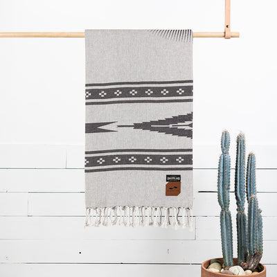Beach blanket with geometric pattern and fringed hem, displayed on wooden bar.