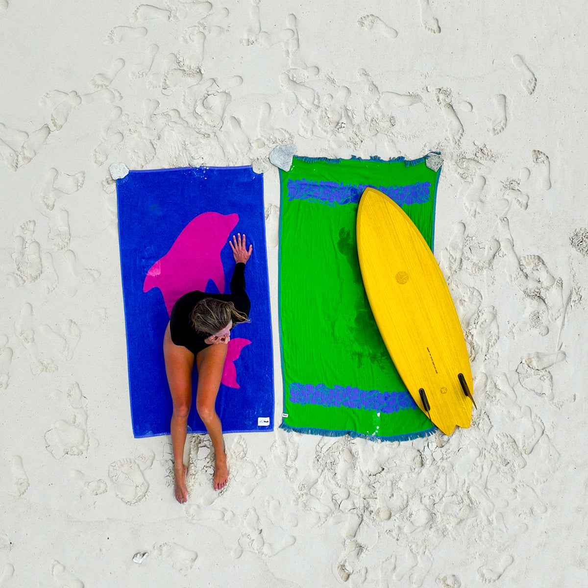 Turkish towel with dolphin design on sand next to green towel and yellow surfboard.