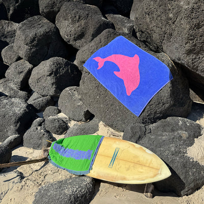 Woven fast-drying towel with pink dolphin design on rocks next to surfboard at beach.