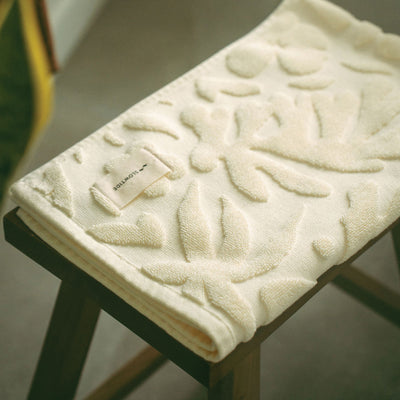 Cream cotton bath towel with raised botanical pattern on a wooden stool.