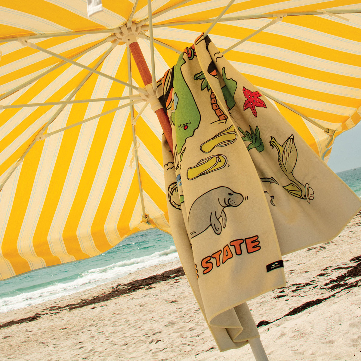 Beach umbrella with yellow stripes and quick-dry towel with Florida print by the ocean.