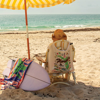 Beach chair under striped umbrella with surfboard, towel, and large tote nearby on sand.