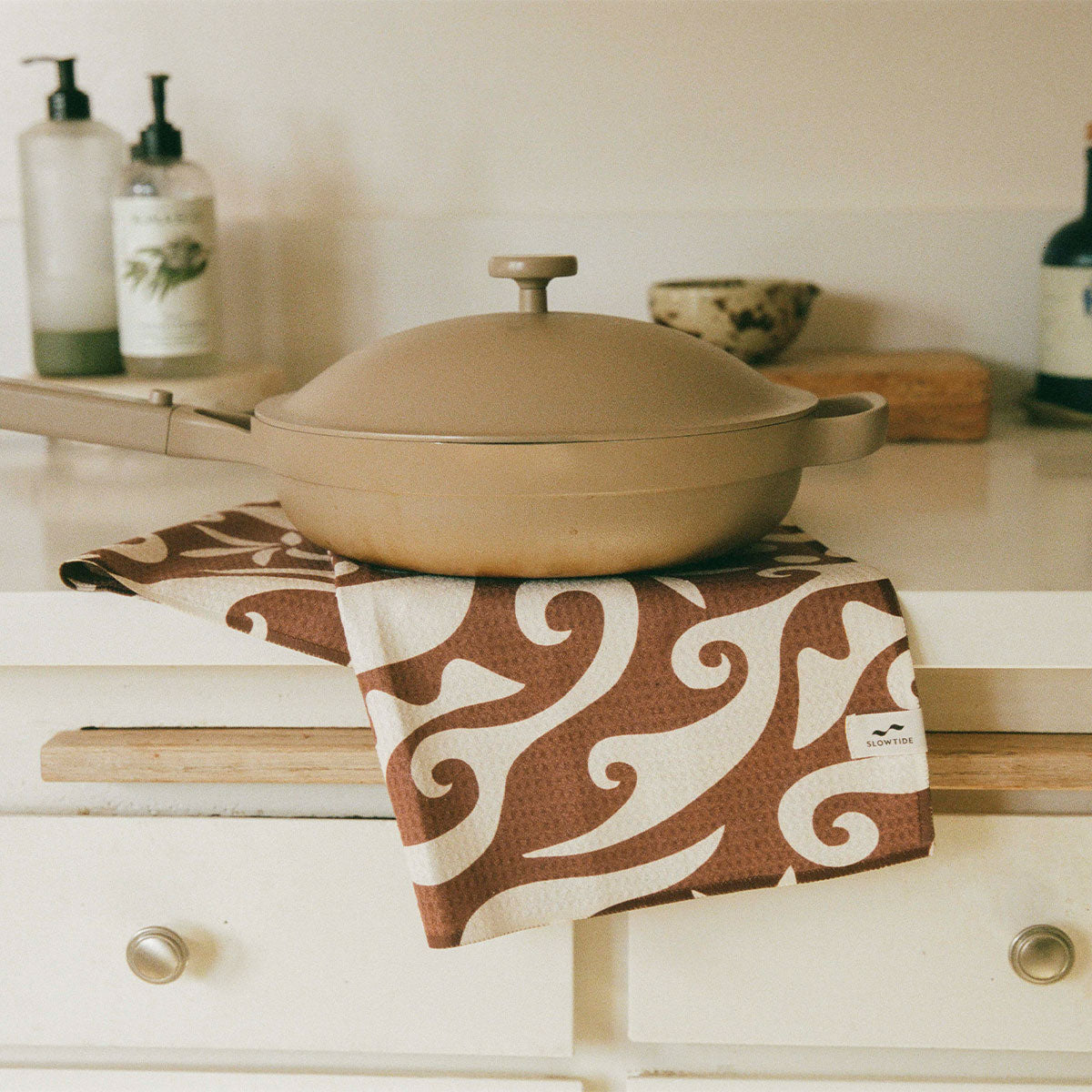 Beige pan with lid on sand-resistant towel on white kitchen counter.
