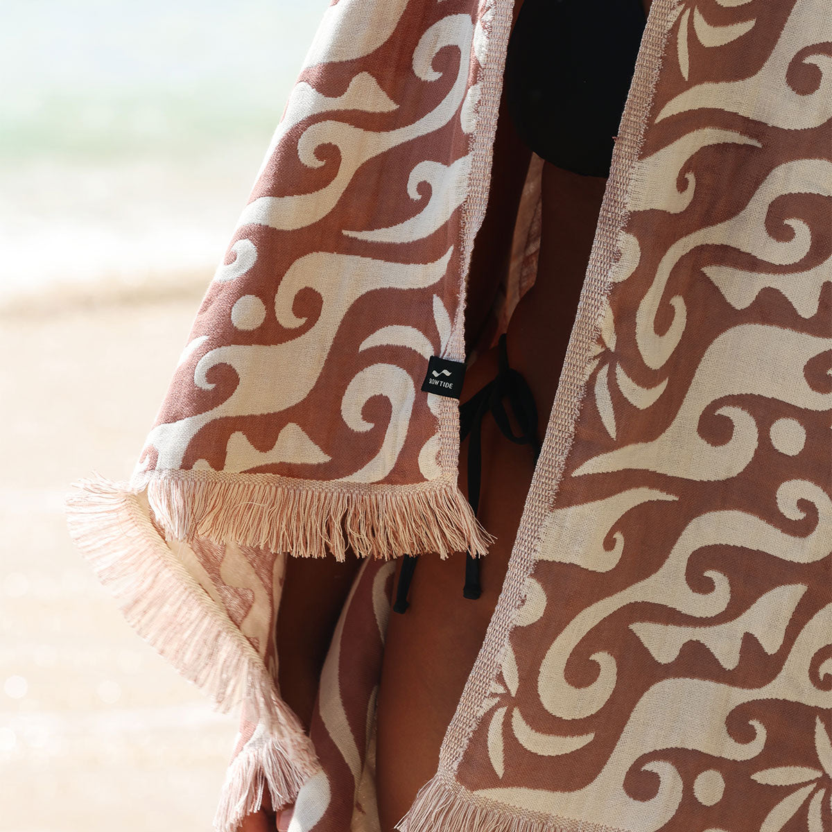 Fringed beach towel with brown and cream wave print on sand.