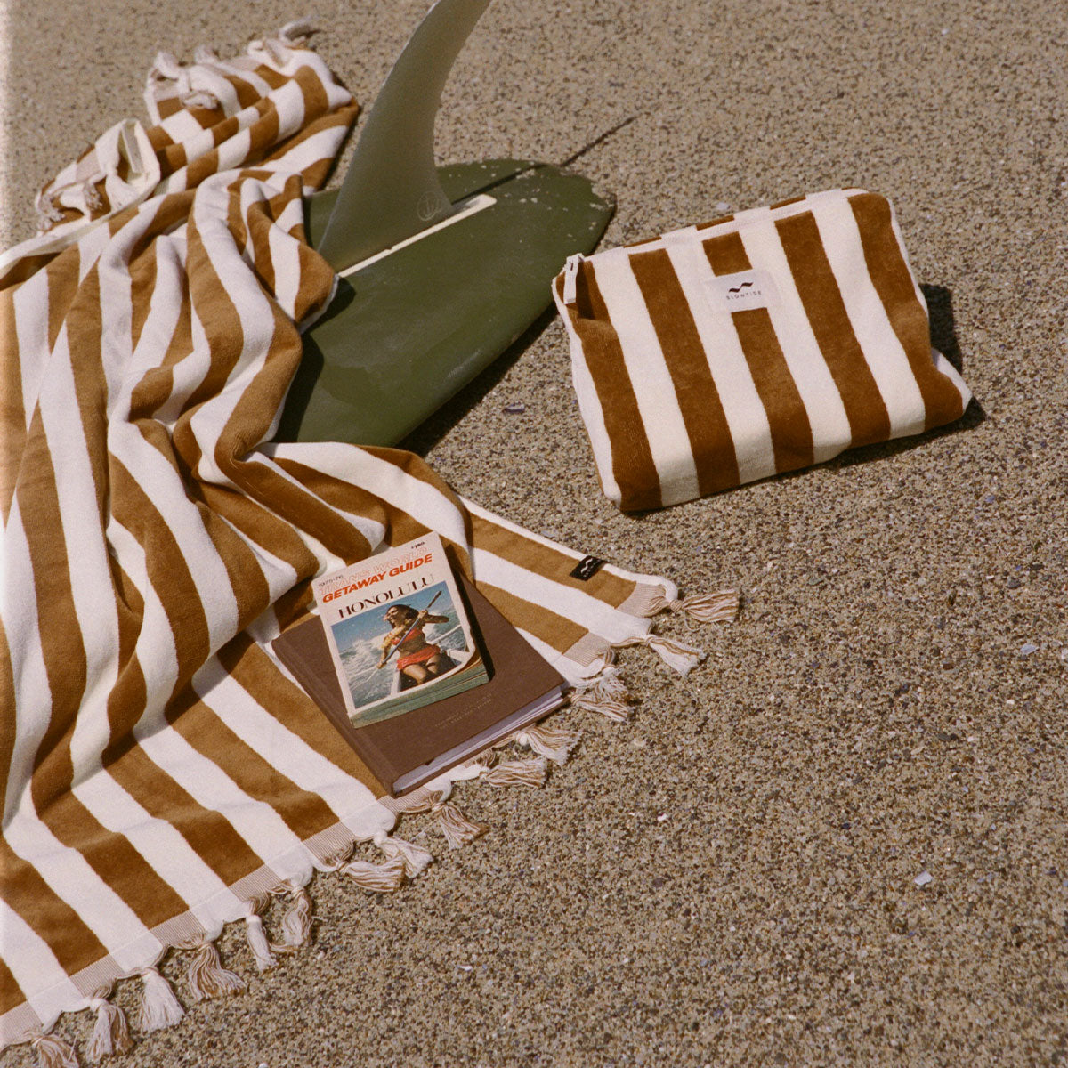 Striped cotton towel with canvas pouch on sand near green surfboard fin.