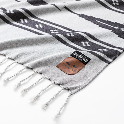 Picnic blanket with black geometric patterns, white fringe, and two corner labels.