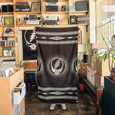 Black woven cotton towel with white lightning skull design in a record store setting.