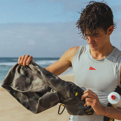Travel towel with camo print pattern shown on a sandy beach near the ocean.