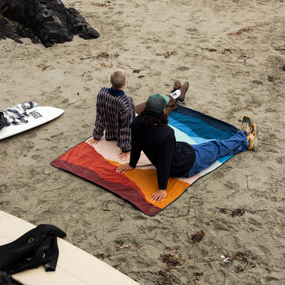 Water-repellent rectangular beach blanket with a multi-colored desert print on sand.