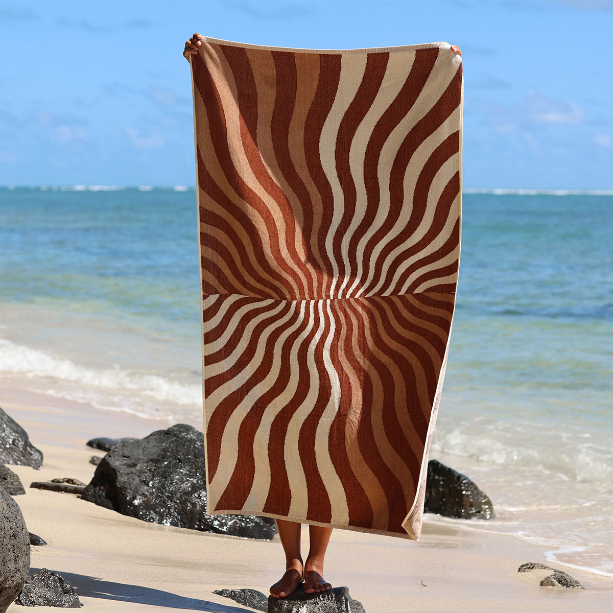 Oversized cotton towel with wavy brown and cream stripes on a sandy beach.