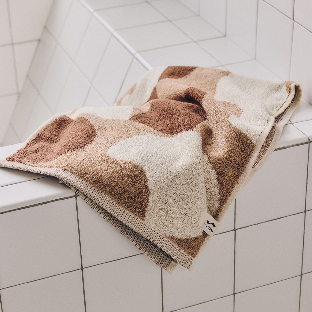 Cotton bath towel with a tan and white abstract pattern draped over a tiled ledge.