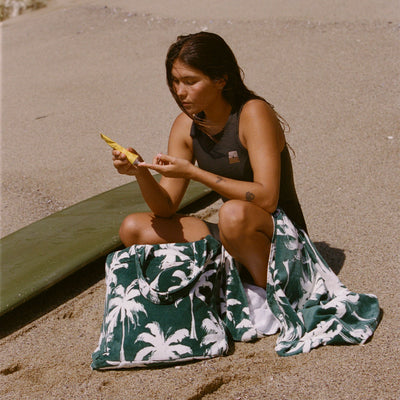 Palm print beach towel and large tote bag on sand next to surfboard.