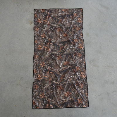 Camo quick-dry towel with brown and green leaf pattern on grey floor.