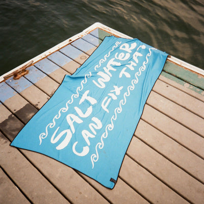 Blue quick-drying towel with SALT WATER CAN FIX THAT! text displayed on a dock.