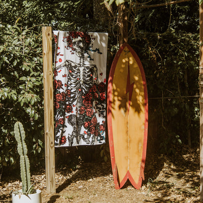 Velour beach towel with skeleton and roses print displayed beside a yellow surfboard outdoors.