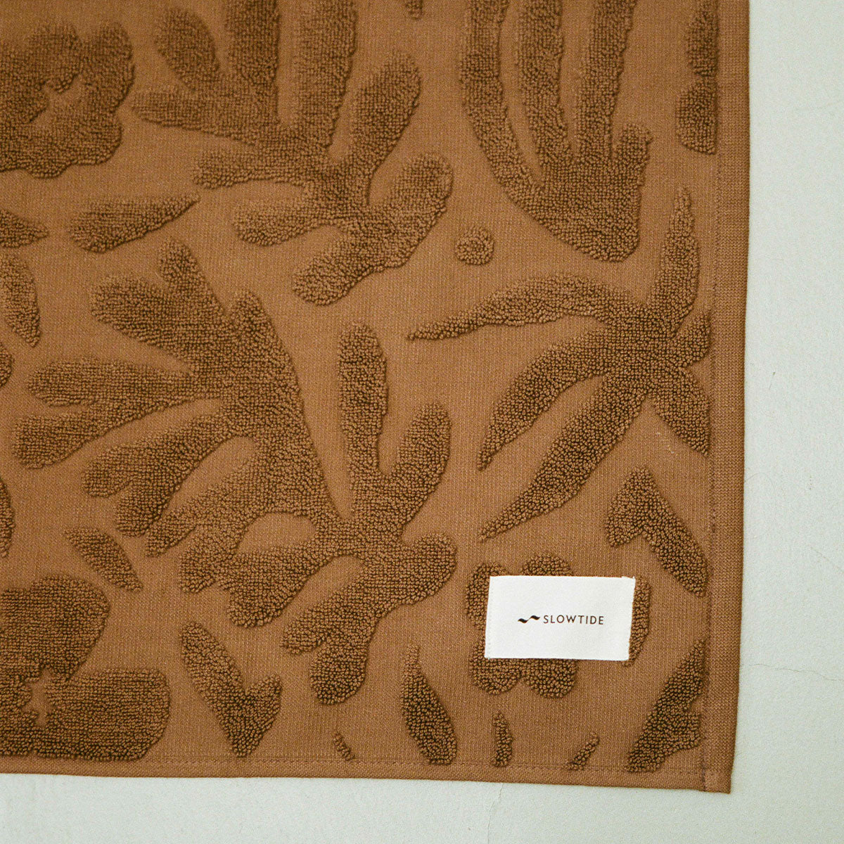 Brown plush towel with textured leaf pattern and small white label.